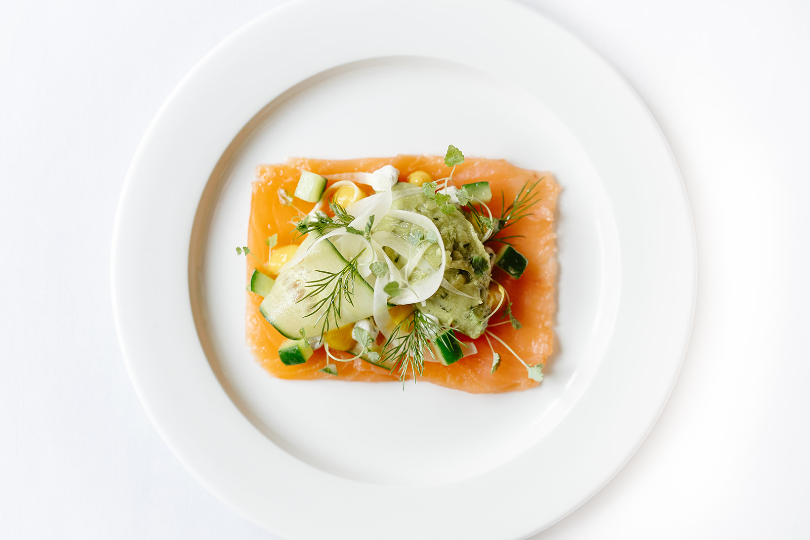 A rectangular slice of smoked salmon topped with fennel, cucumber ribbons, and herbs, served on a white plate.
