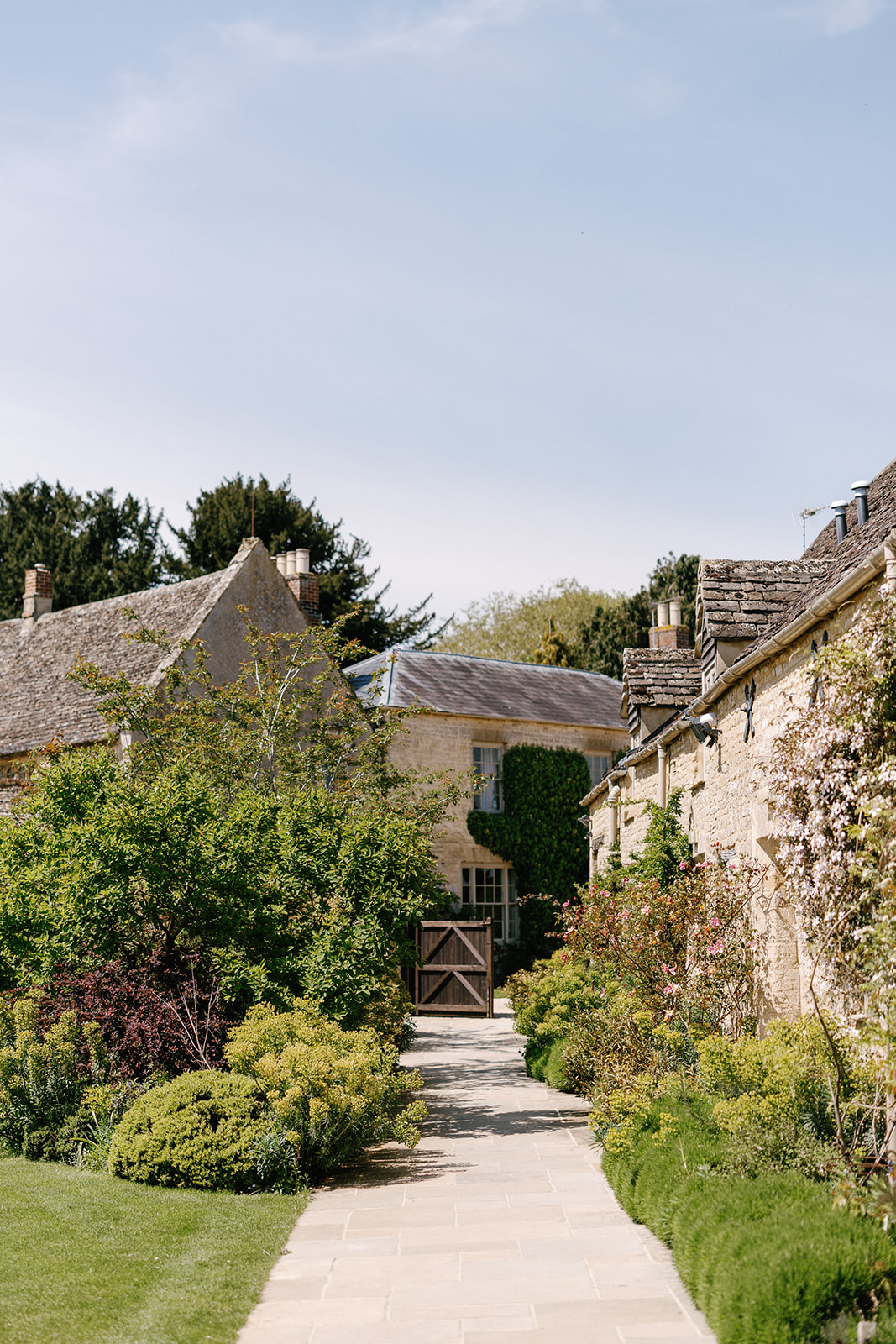 A stone pathway leads between two ivy-covered stone buildings, surrounded by lush garden greenery under a clear blue sky.