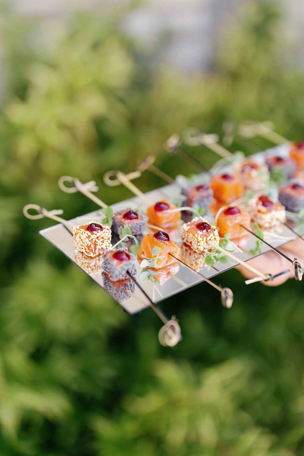 A close-up of several skewered bite-sized appetizers with garnish, arranged neatly on a reflective tray, held outdoors against a blurred green background.